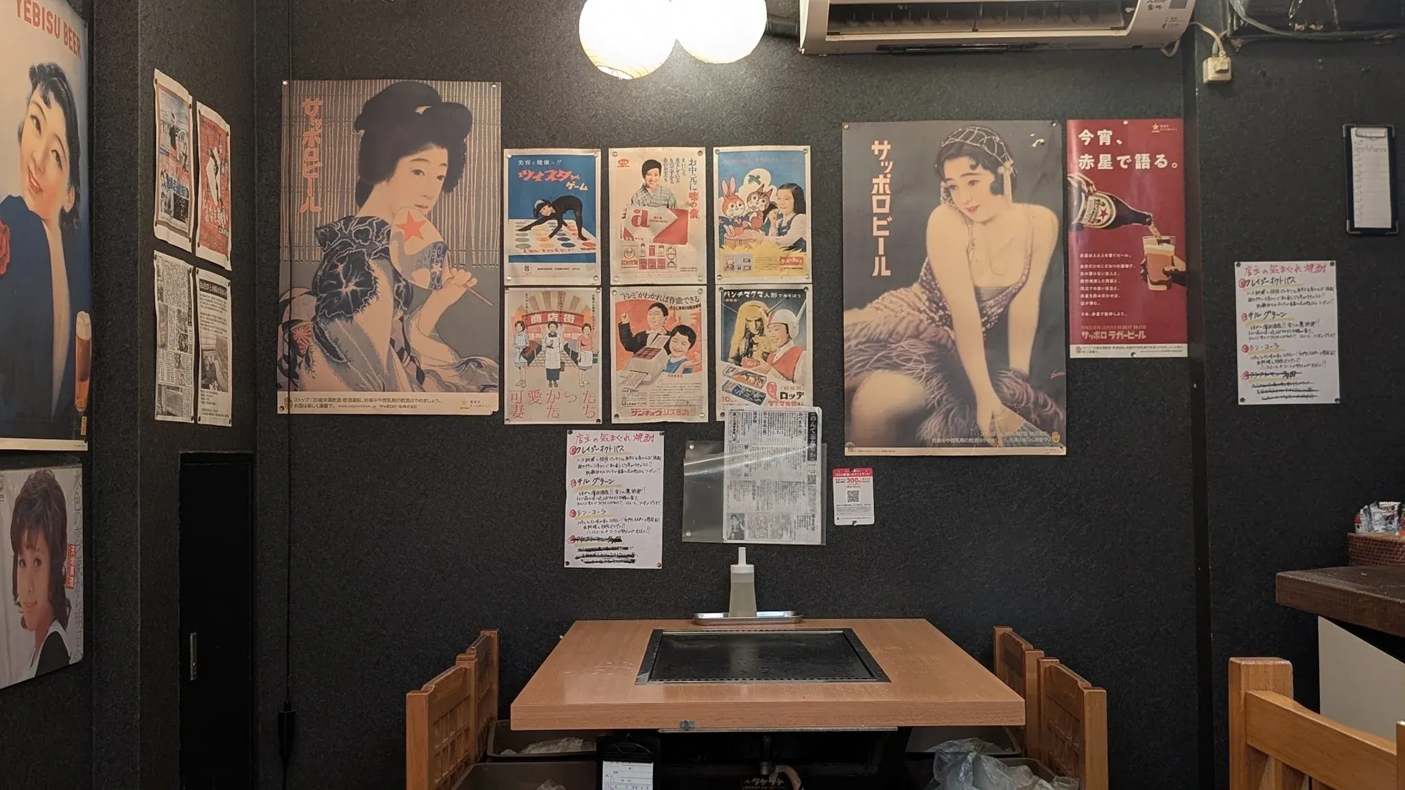 A small restaurant table with a built‑in grill, set against a wall decorated with Shōwa‑era Japanese posters.