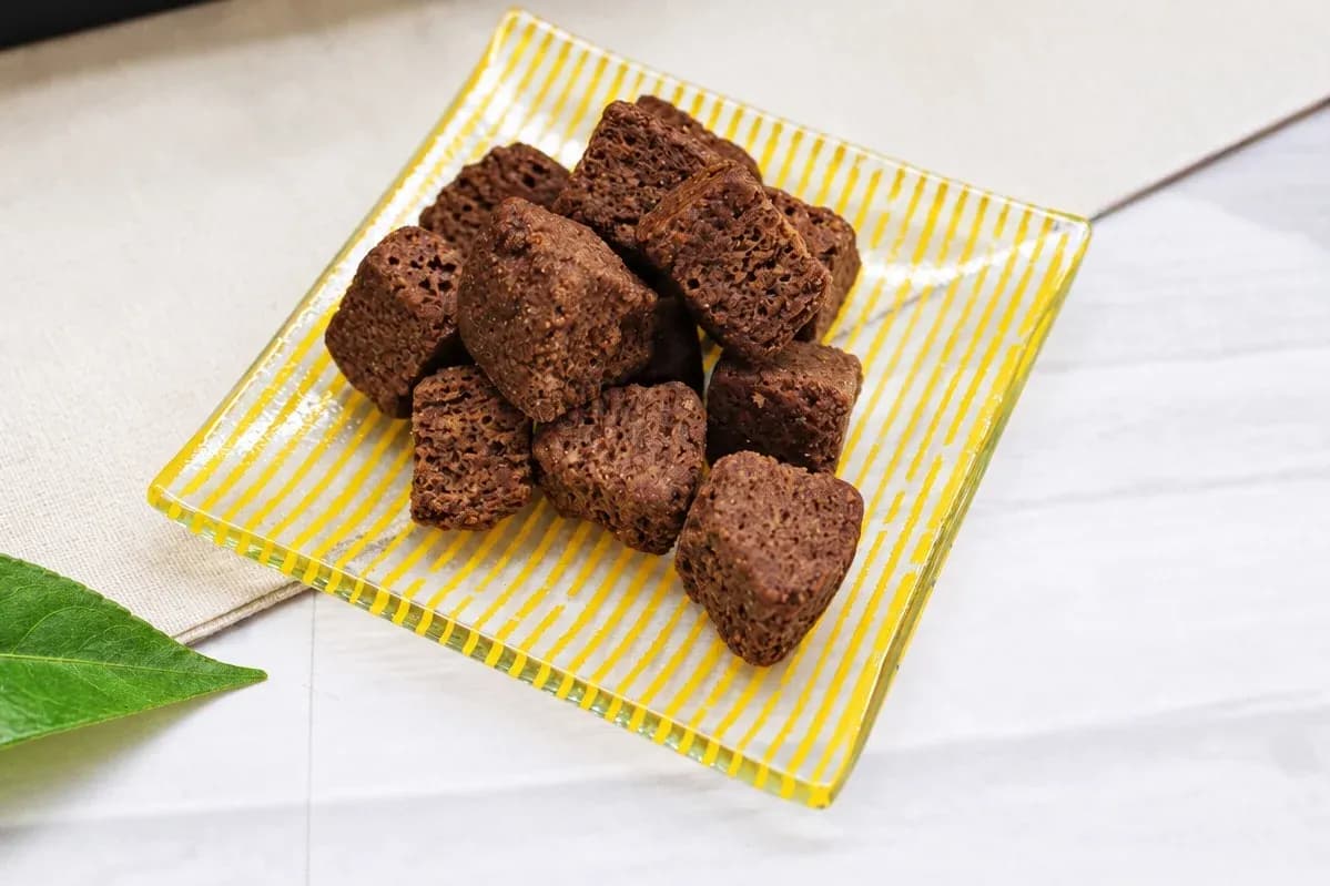 A yellow striped plate holding several cube-shaped pieces of chocolate rusk with a rich brown color and crisp texture.