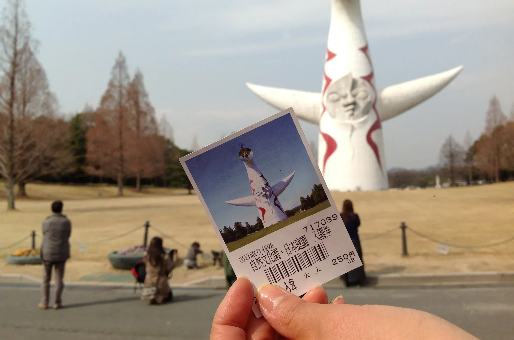 万博記念公園の入場券を手に持ち、背景に太陽の塔がそびえている。