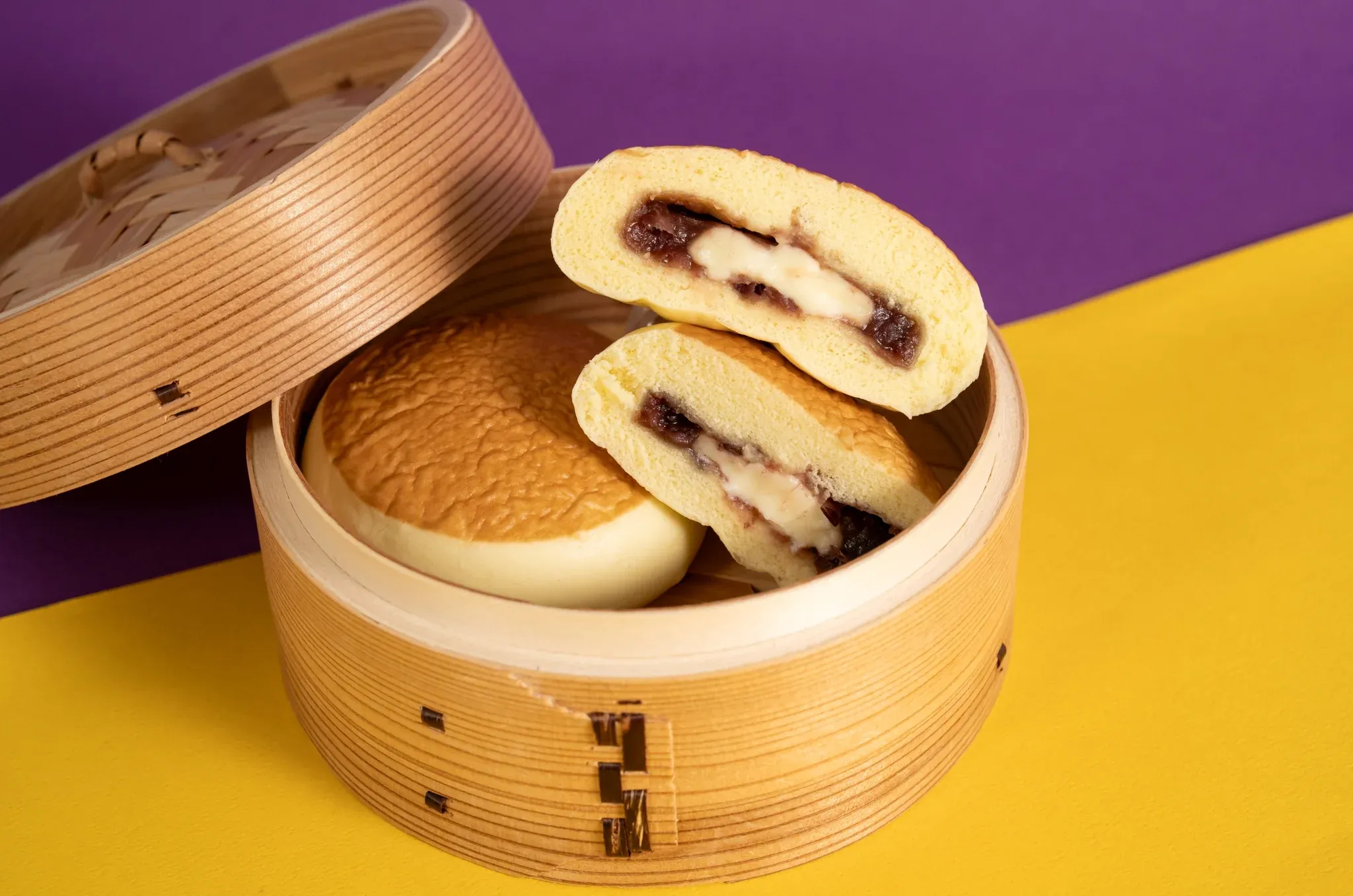 紫と黄色の背景に、竹製せいろの中で丸いホットケーキ風まんじゅうが並ぶ。半分に切られたまんじゅうから、あんことバタークリームの層が見える。