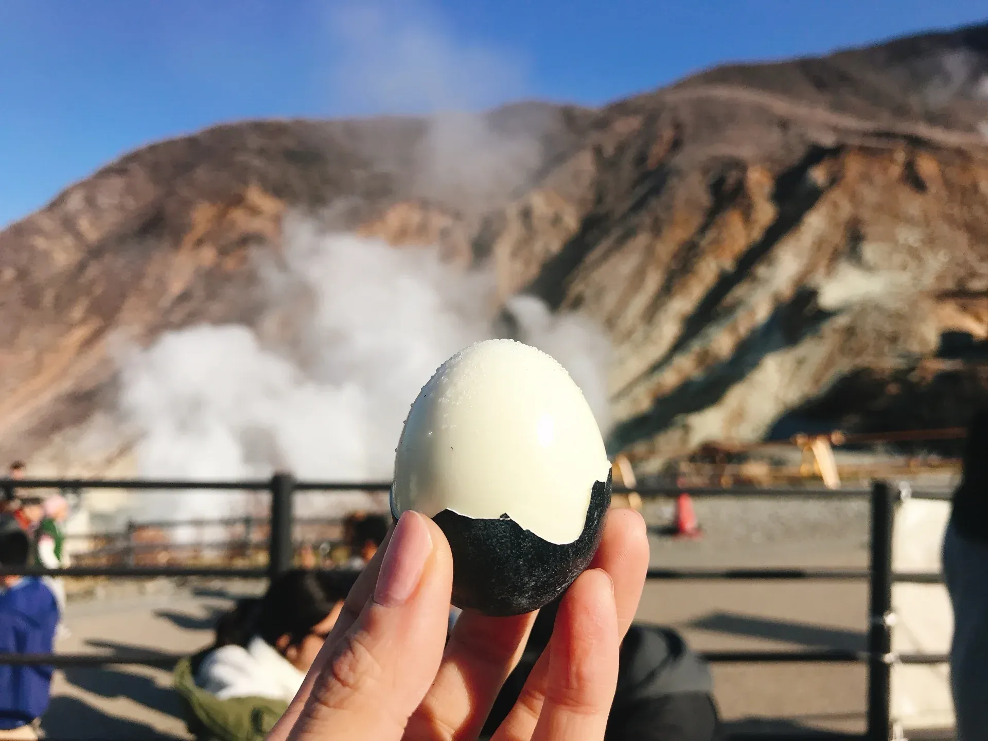 A hand holds one of Owakudani’s black eggs with volcanic steam and rocky slopes in the background under a clear blue sky.