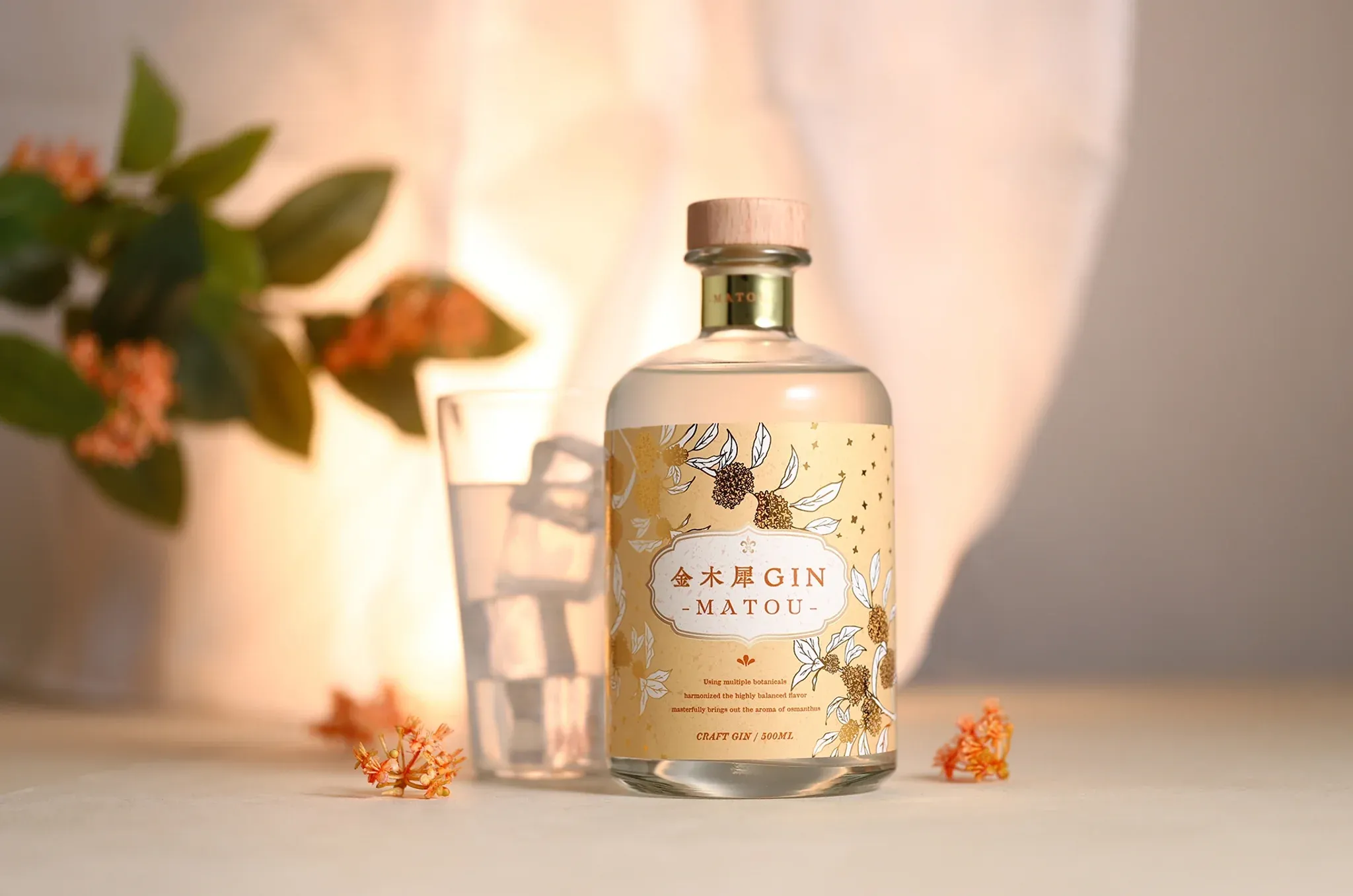 氷水の入ったグラスの隣に、金木犀の花が添えられた金木犀GIN -MATOU-のボトルが置かれている。背景には緑の葉と柔らかな光が広がる。