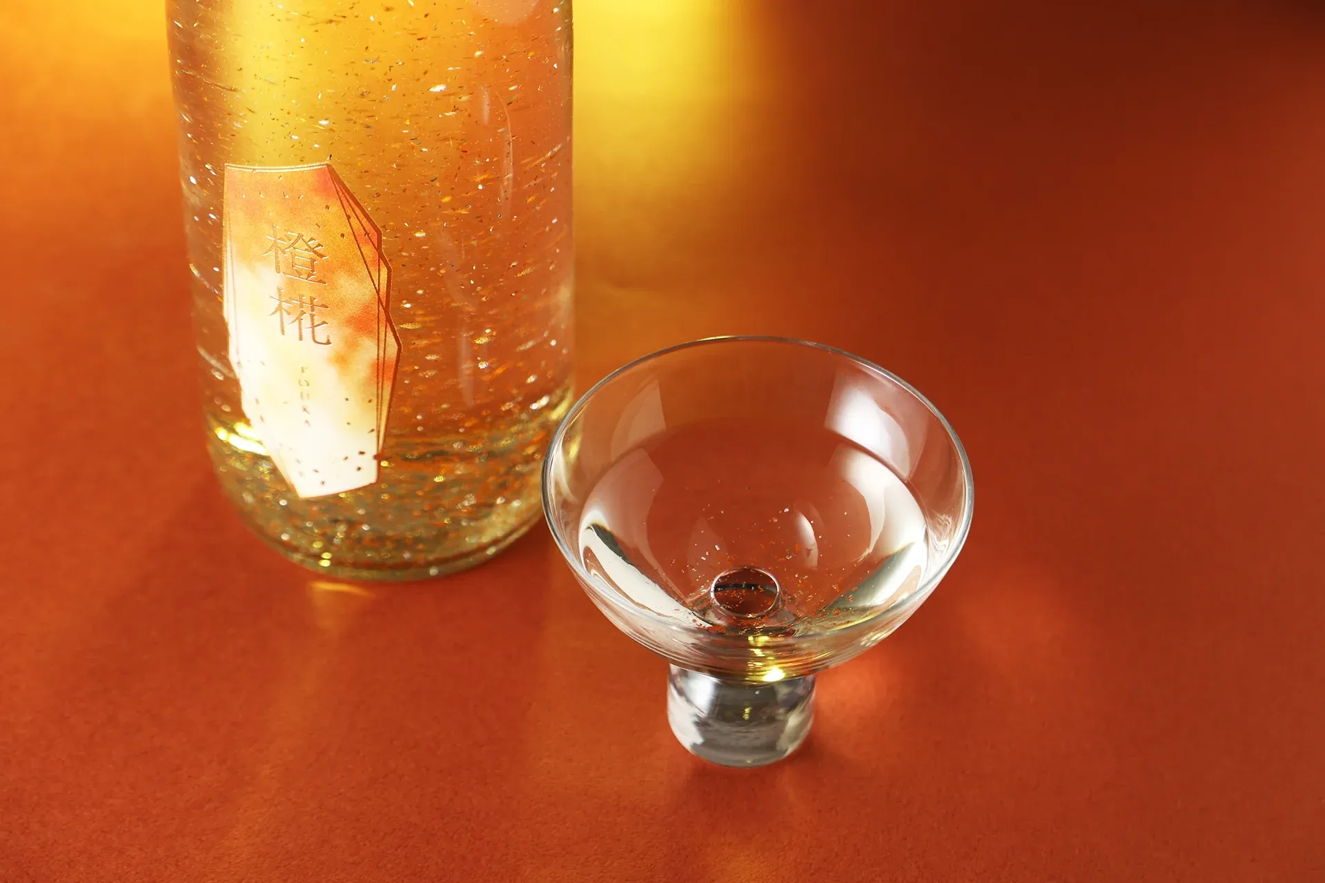 オレンジの背景に置かれた橙椛の酒瓶と杯。液体の中で金箔がきらめく。