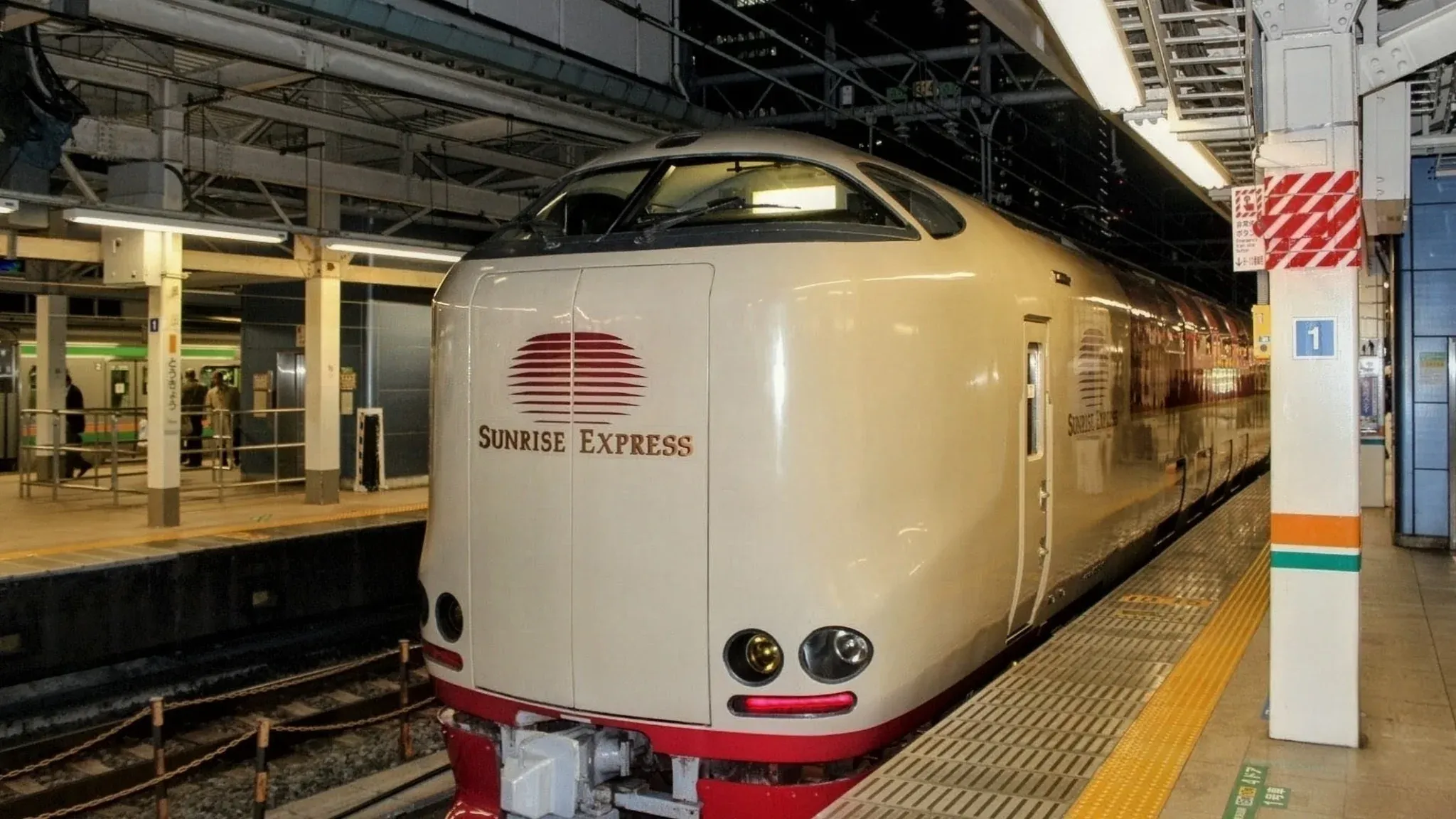 駅のホームに停車するベージュ色のサンセットエクスプレス。丸みを帯びた先頭と赤い太陽のロゴが印象的。